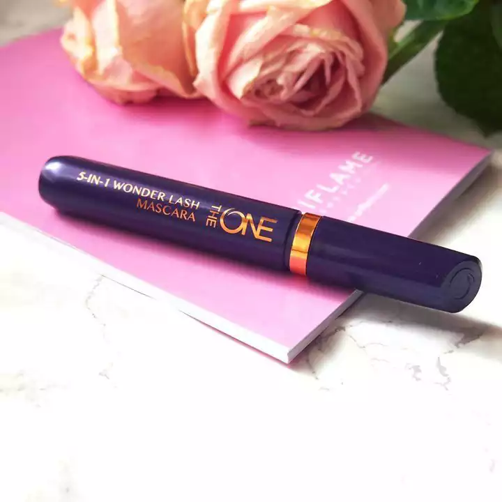 یک تیوب ریمل 5 در 1 Wonder Lash از برند The One، که روی یک کتابچه صورتی با یک گل رز صورتی در پس زمینه قرار گرفته است. تیوب ریمل به رنگ بنفش تیره با حروف طلایی و یک نوار نارنجی است که روی یک سطح مرمری قرار گرفته است.